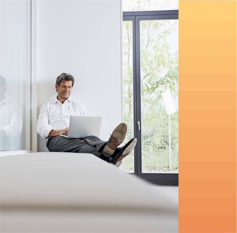 Man reclines on a bench while working from his laptop in an office building