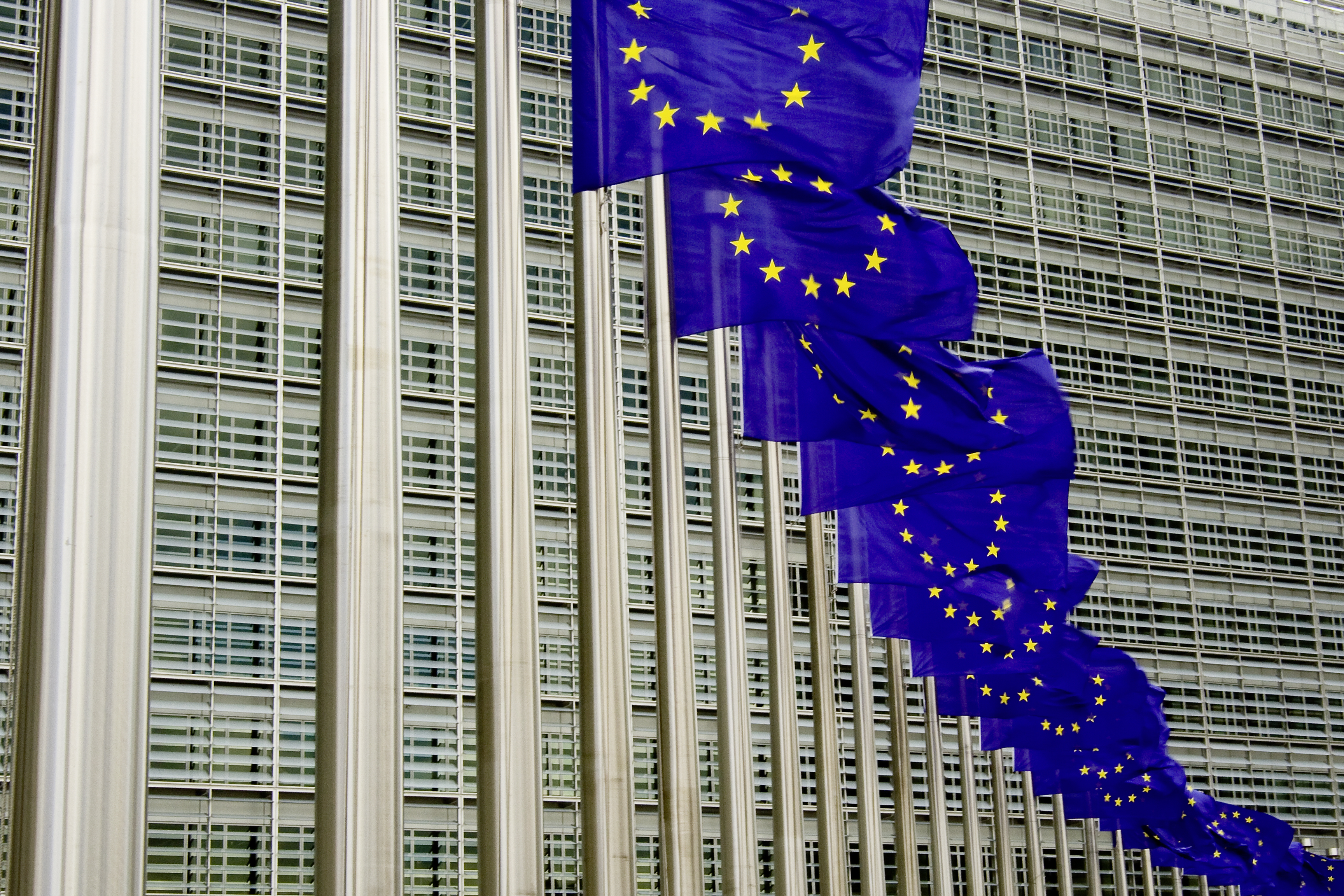 This image shows a row of blue EU flags with stars flying in front of the European Commission and Parliament buildings in Brussels.